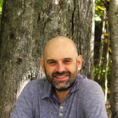 A white man with a shaved head and facial hair smiles while sitting in front of a tree