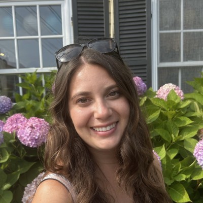 A brunette woman with sunglasses on top of her head smiles while standing in front of a bush of pink hydrangeas
