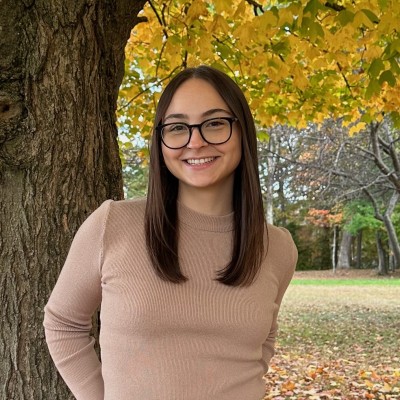 A white woman with straight brown hair and wearing glasses and a beige sweater smiles in front of an autumn tree