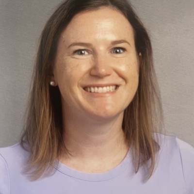 A white woman with shoulder-length brown hair and wearing a pale purple shirt smiles
